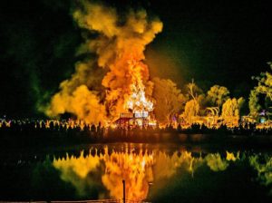 the temple of spirals is burning from across the lake in orange and yellow smoke