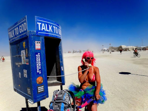 Woman with pink hair and a tutu talking on the phone at a phonebooth with the sign "Talk to God"