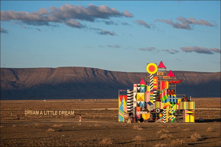 The desert, with two large artworks in the distance: the letters "Dream a little dream" on the left, and a colourful, geometrically inspired castle in strong primary colours and black and white on the left.