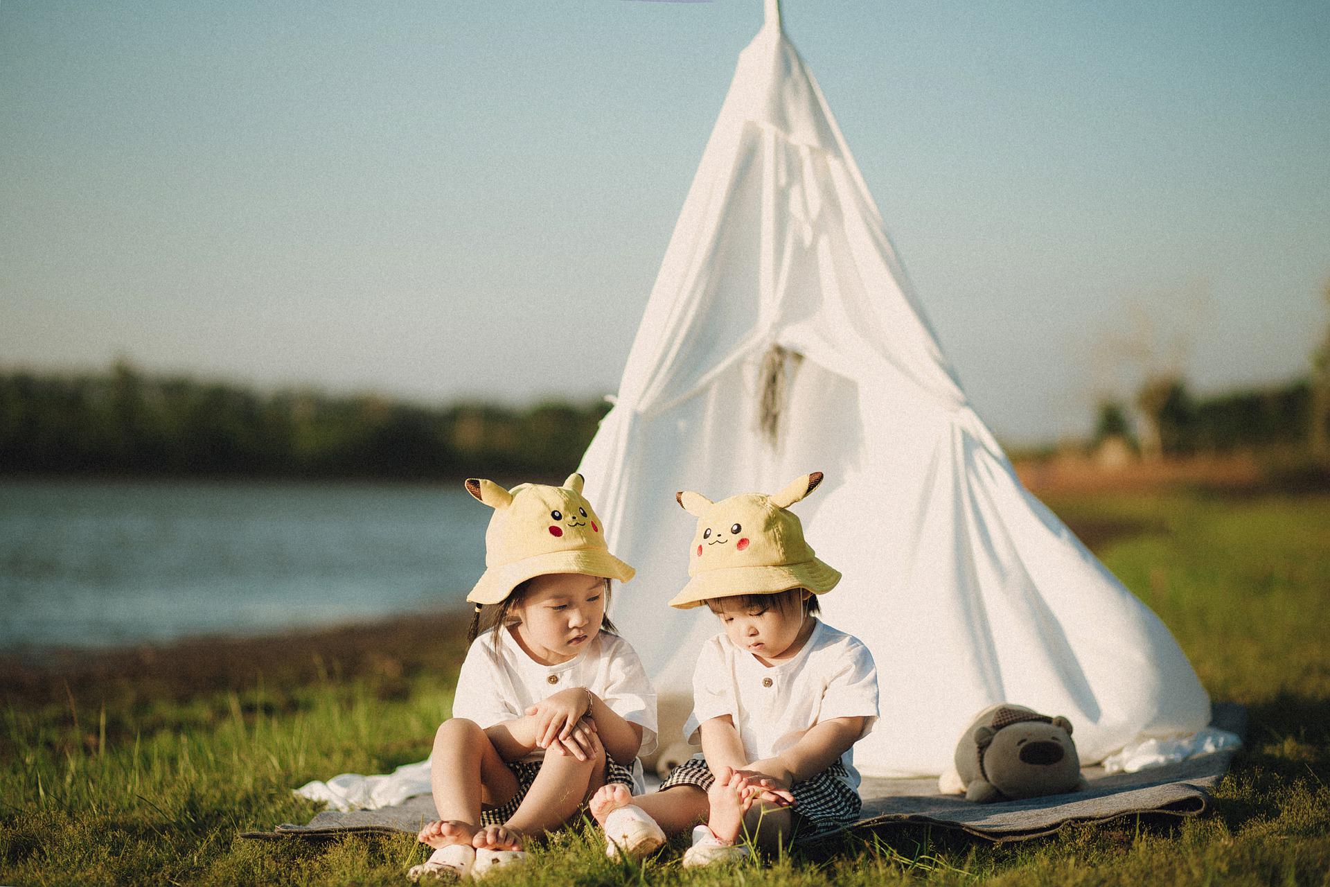 Two children sit in front of a tee-pee style tent with a body of water in the background
