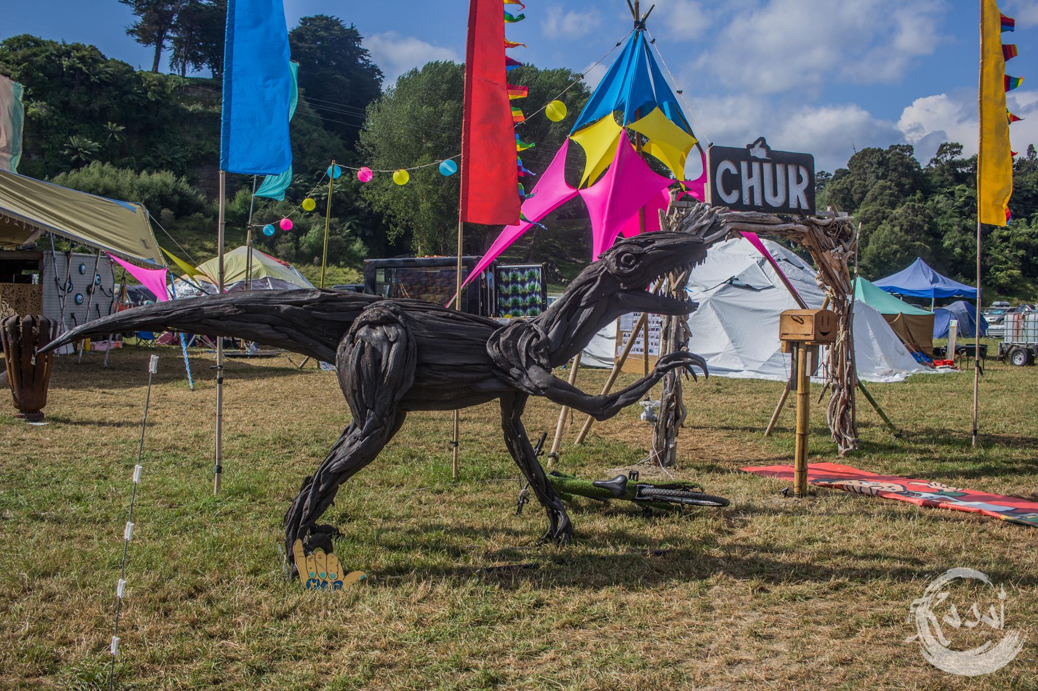 A dinosaur art work sits in front of a vibrantly coloured theme camp on the Paddock