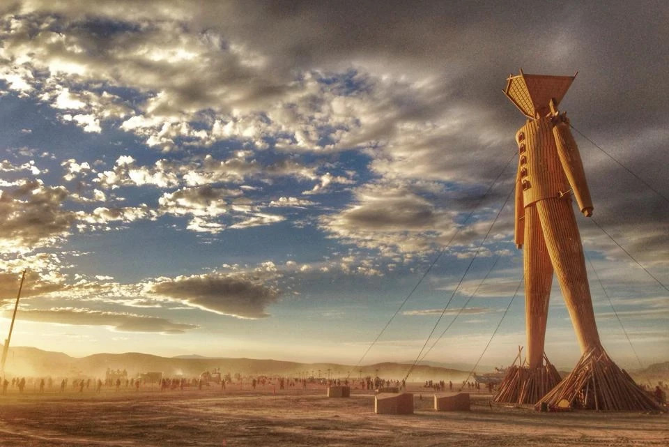 The Man stands tall in the desert with a swirl of clouds against a fading blue sky