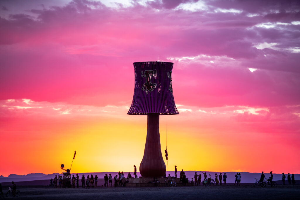 A giant lamp art piece with people swinging from it stands in front of a purple, pink, orange and yellow sky