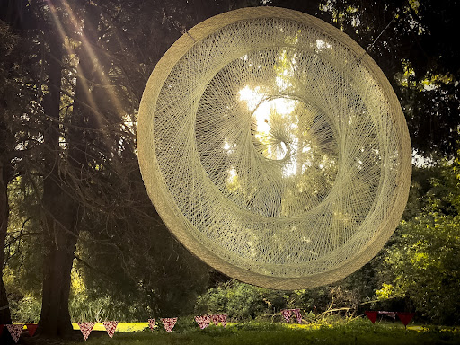 A beige woven dreamcatcher-like tapestry hangs in the forest with dappled sunlight shining through