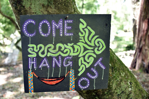A black sign attached to a bent tree branch reads "Come hand out" in purple letters, with a swirly green arrow pointing right and a red hammock underneath