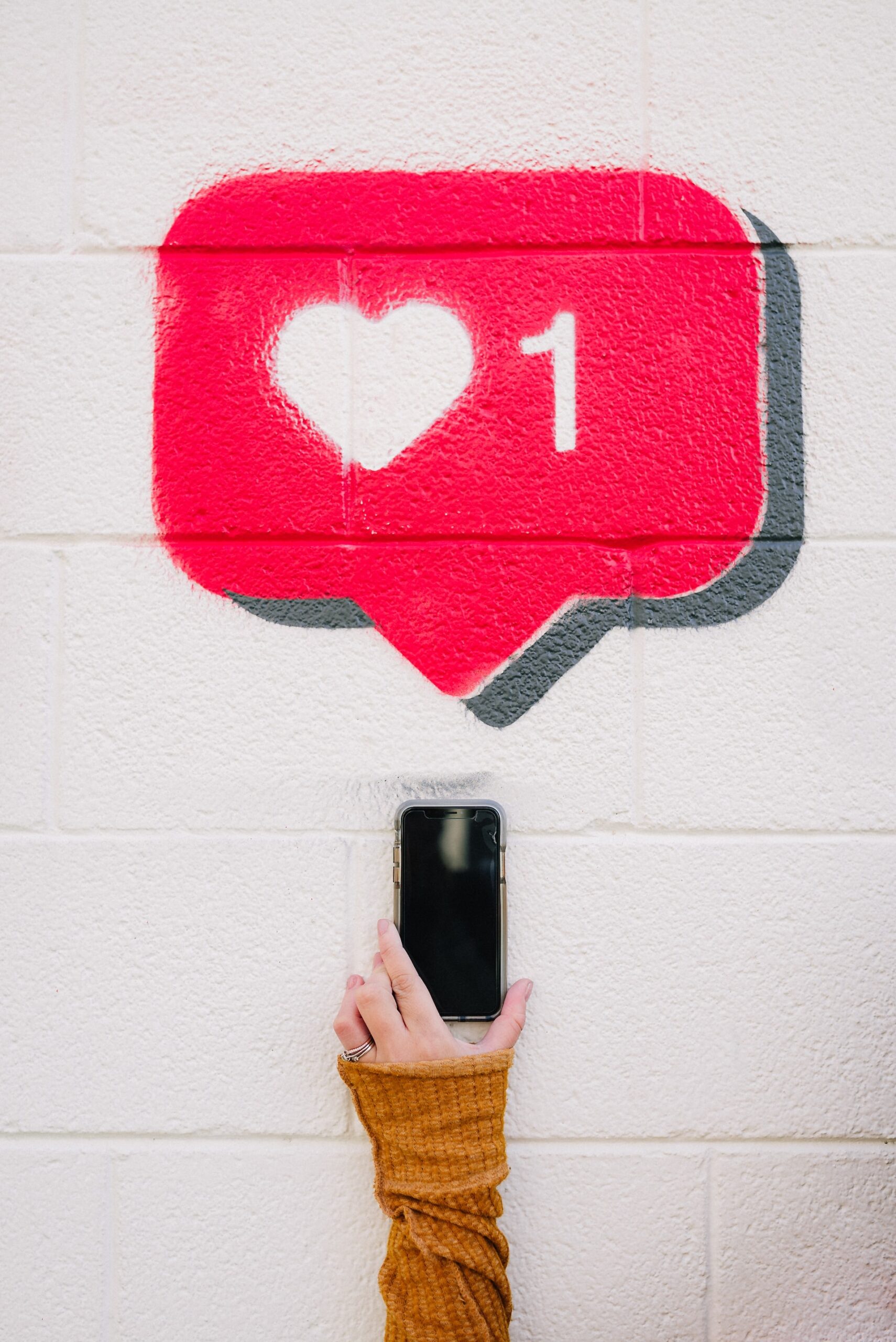 A hand holding a cell phone reaches up against a white brick wall towards a red speech bubble with a white heart and the number 1 inside it