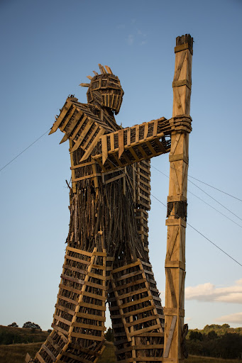 A wooden structure (the Effigy) of a large robot holding a stick stands tall with bright blue sky in the background