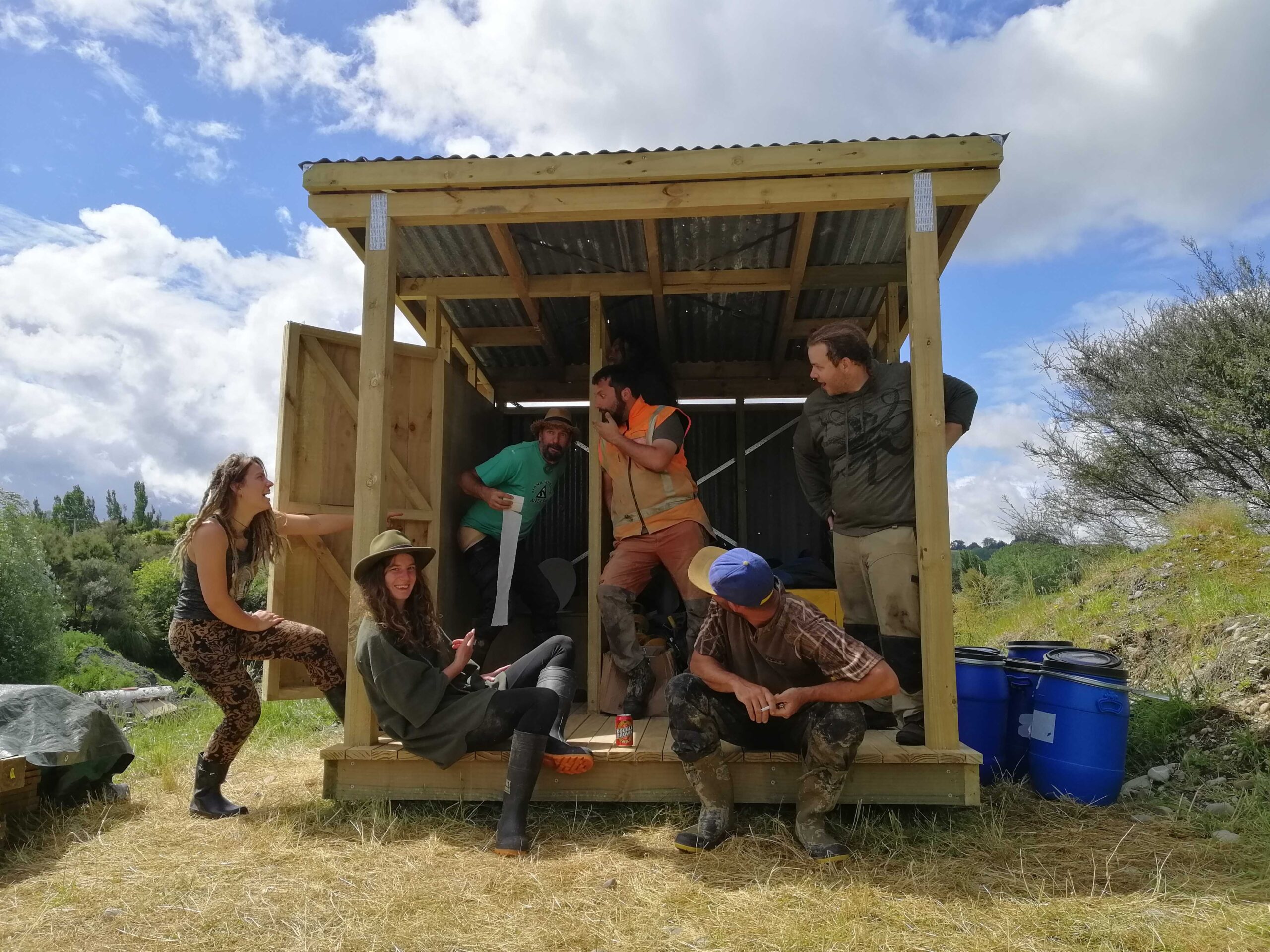 Six volunteers stand in front of a partially built wooden composting toilet building pulling silly poses
