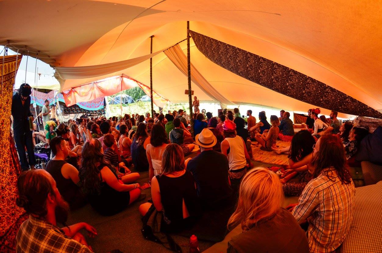 A large group of people sit under a marquee facing a single person in the distance