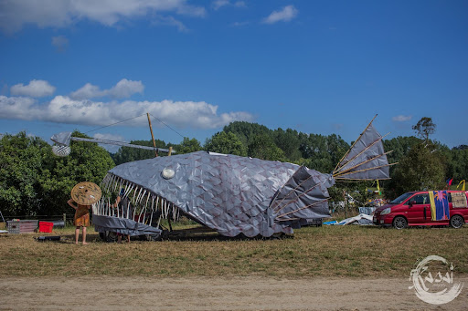 An Art car in the shape of a giant angler fish, with greyish skin and large sharp teeth sits on the paddock