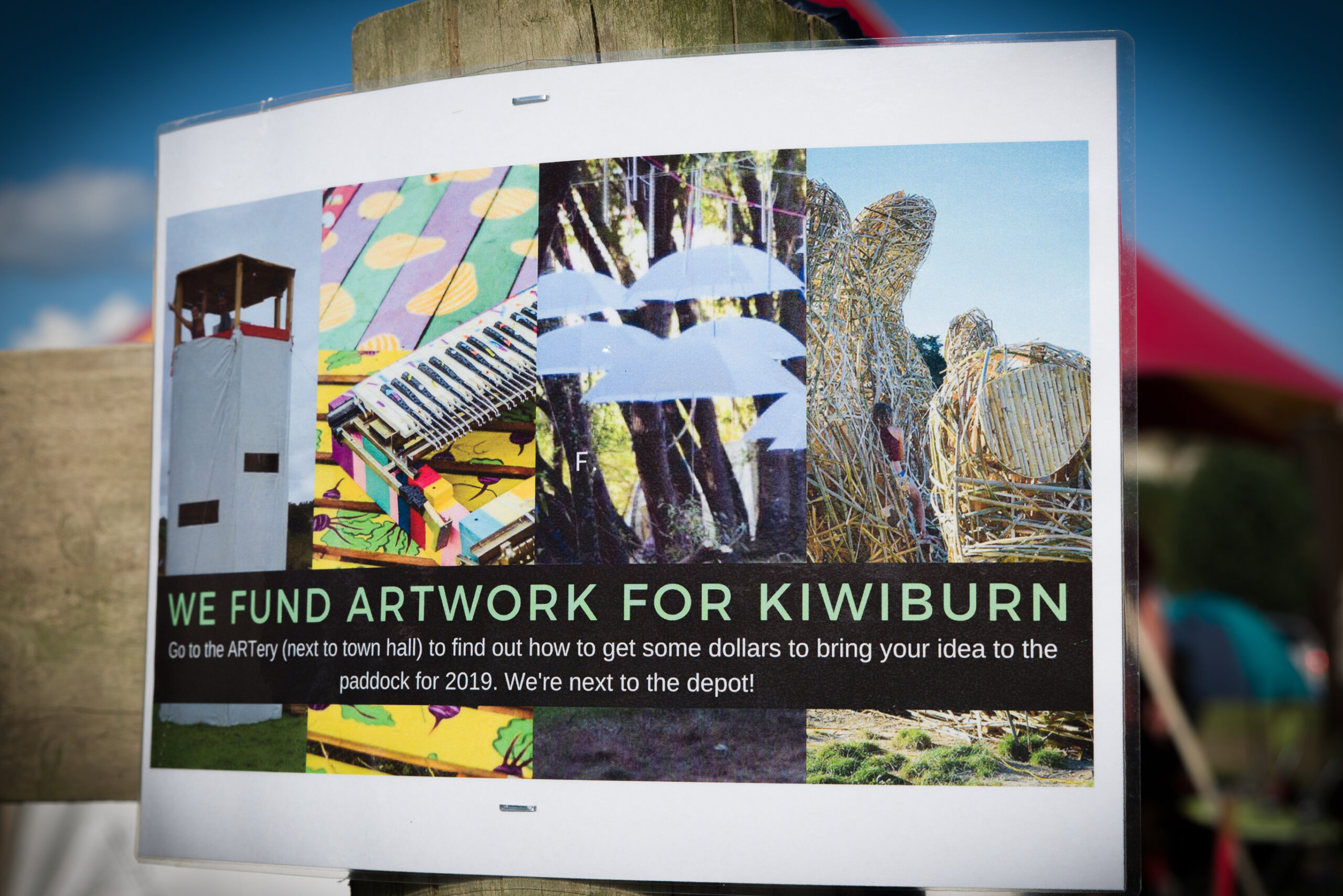 A poster stapled to a wooden post displaying various artwork from previous burns with the words 'we fund artwork for kiwiburn' boldly written at the bottom