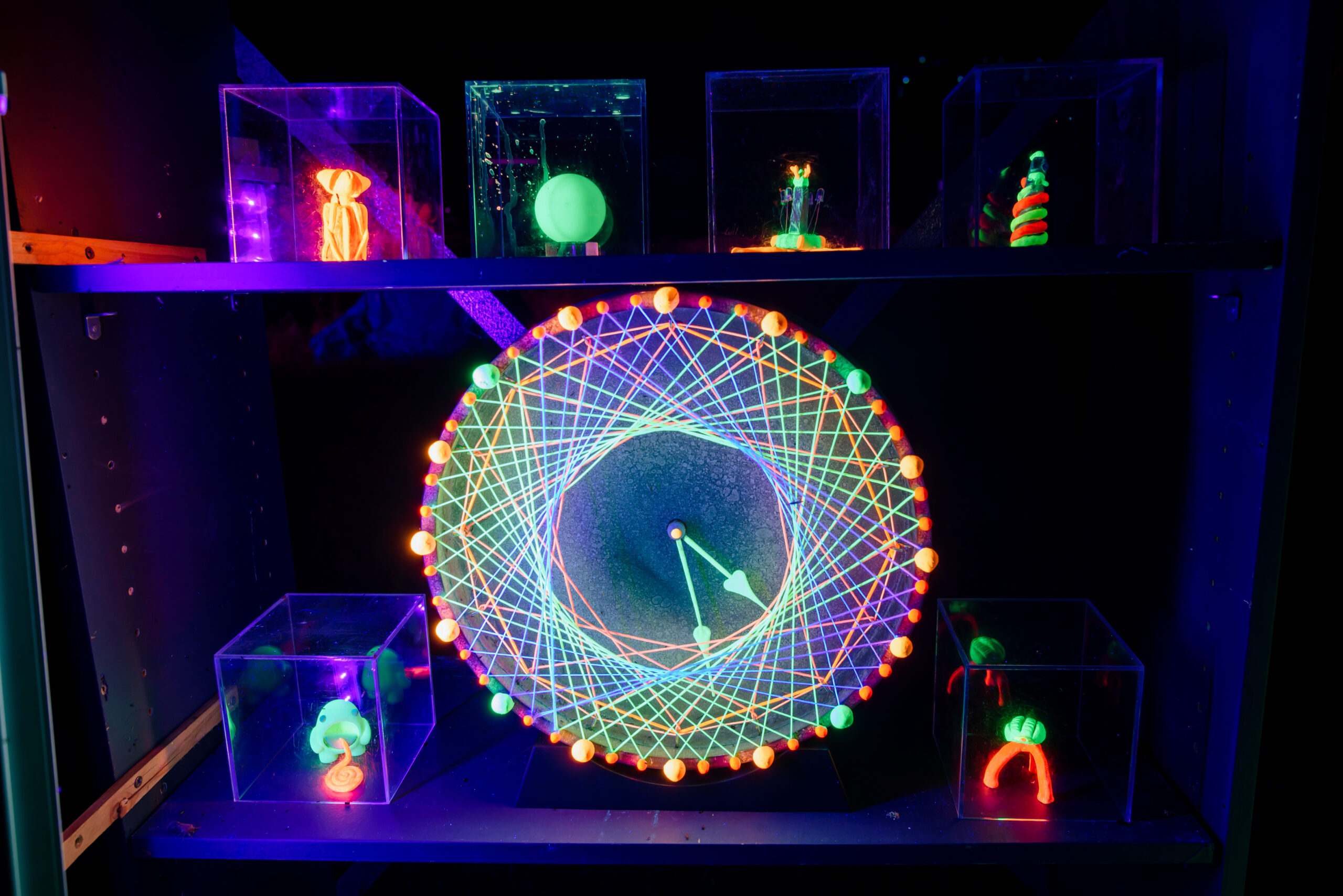 A bright UV glow, mandala style clock sits in the centre of a shelf, surrounded by small glowing random objects in clear boxes