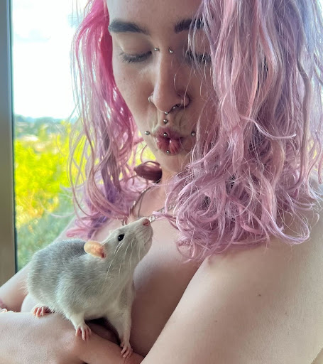 A person with bright pink hair blowing a kiss to a white rat sitting on their arms