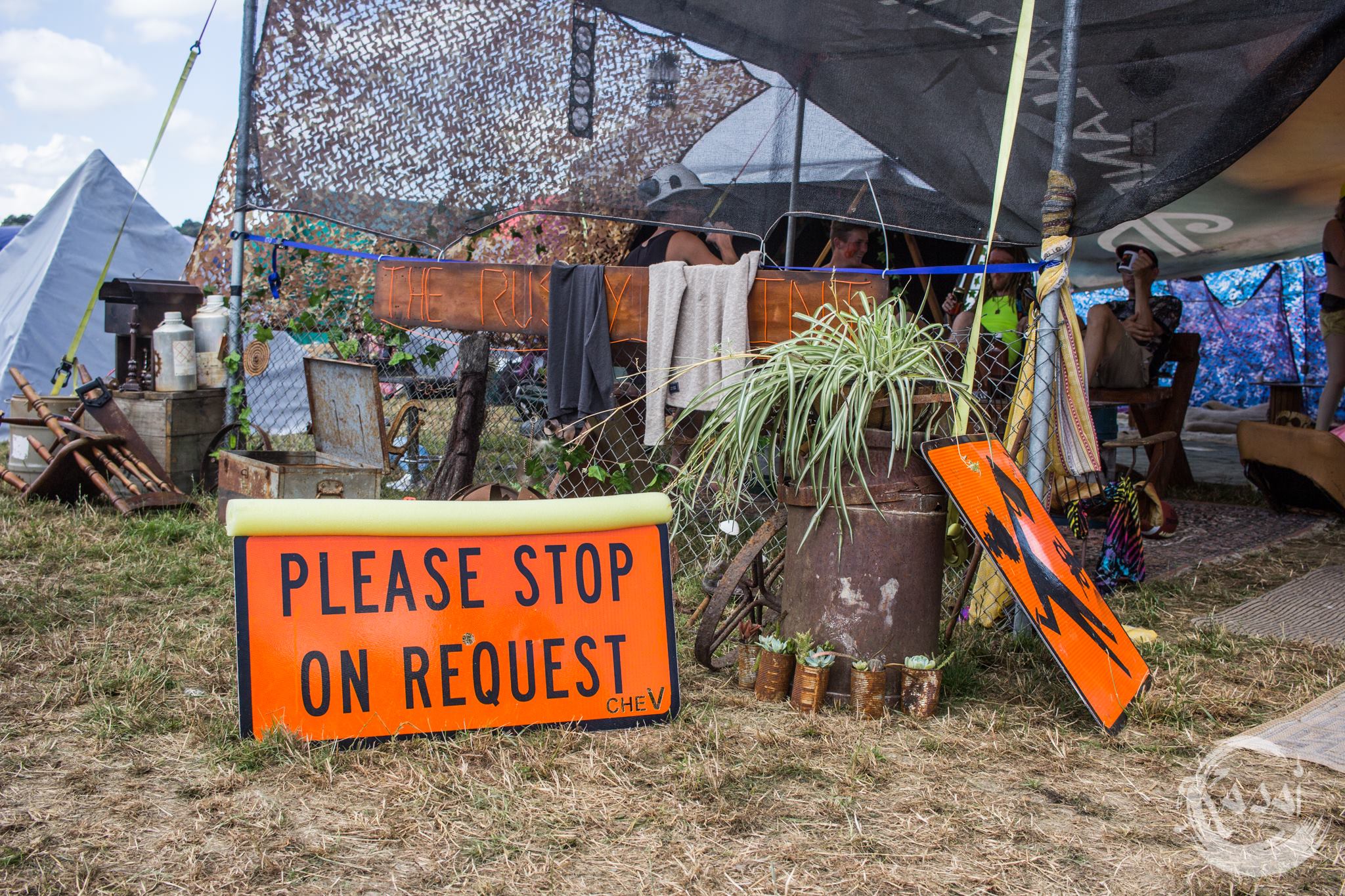 a bright orange sign with bold black lettering saying 'please stop on request' sits in front of a theme camp