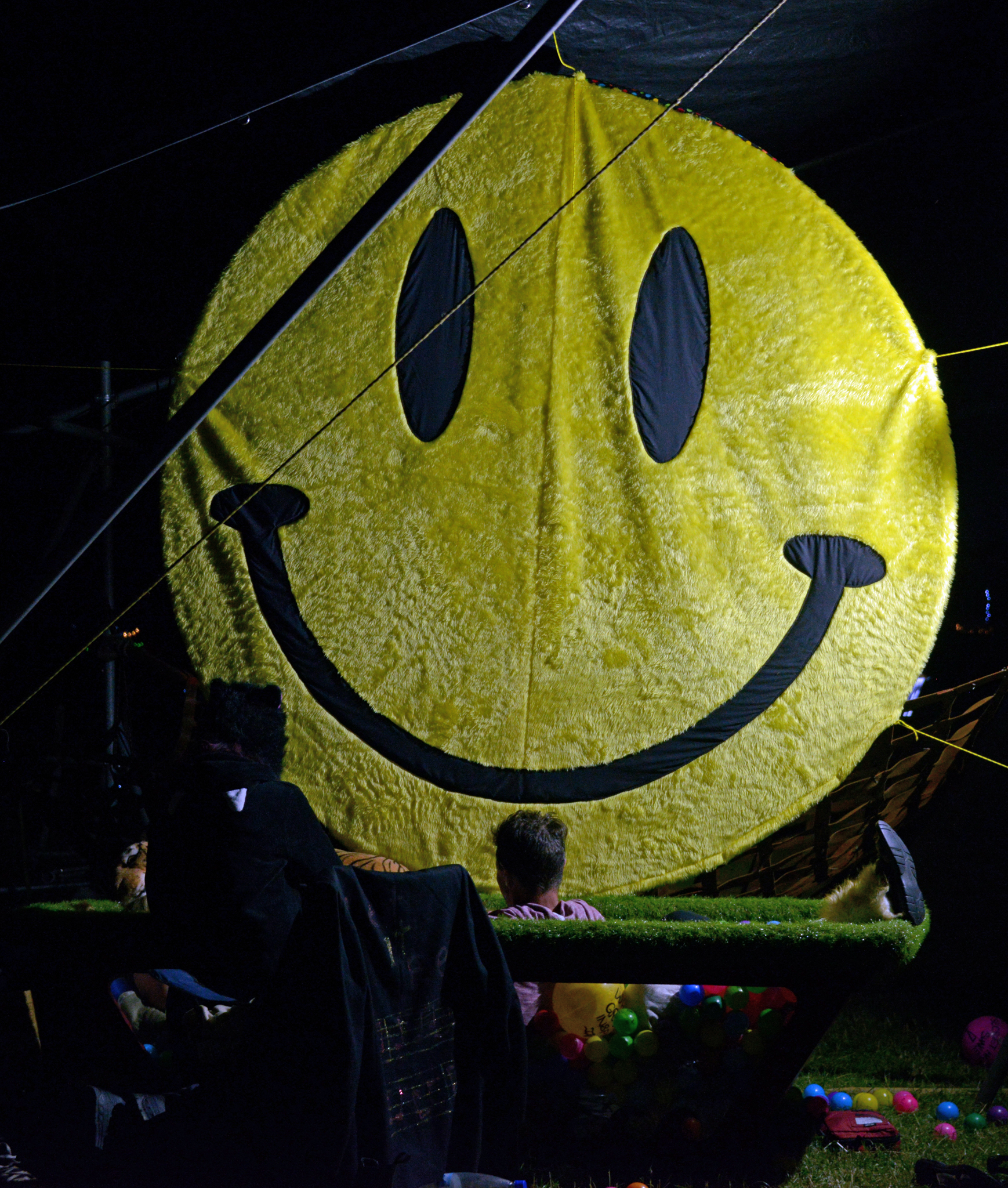a giant yellow smiley face sits against a black backdrop with a pool filled with colourful plastic balls in front of it