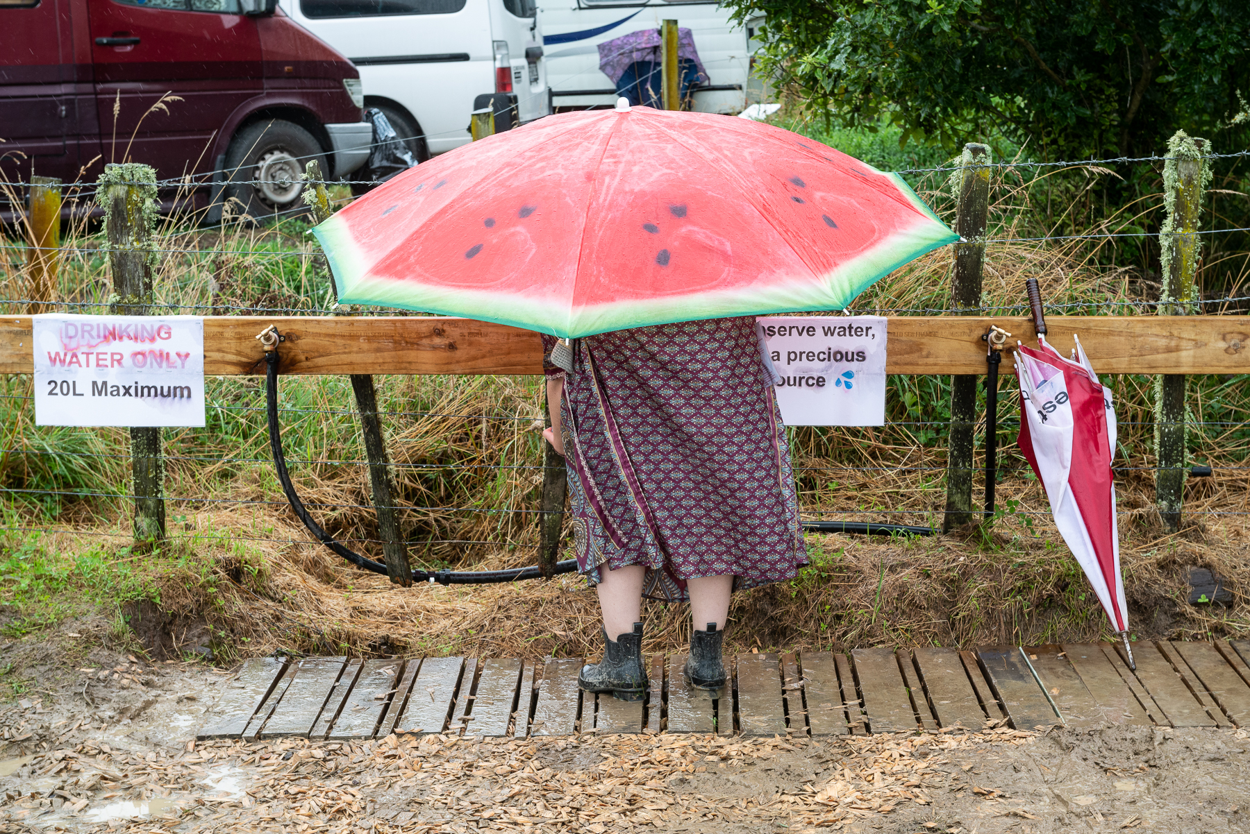 a person stands at the water filling station wearing gumboots and a long dress, holding a bright red watermelon umbrella