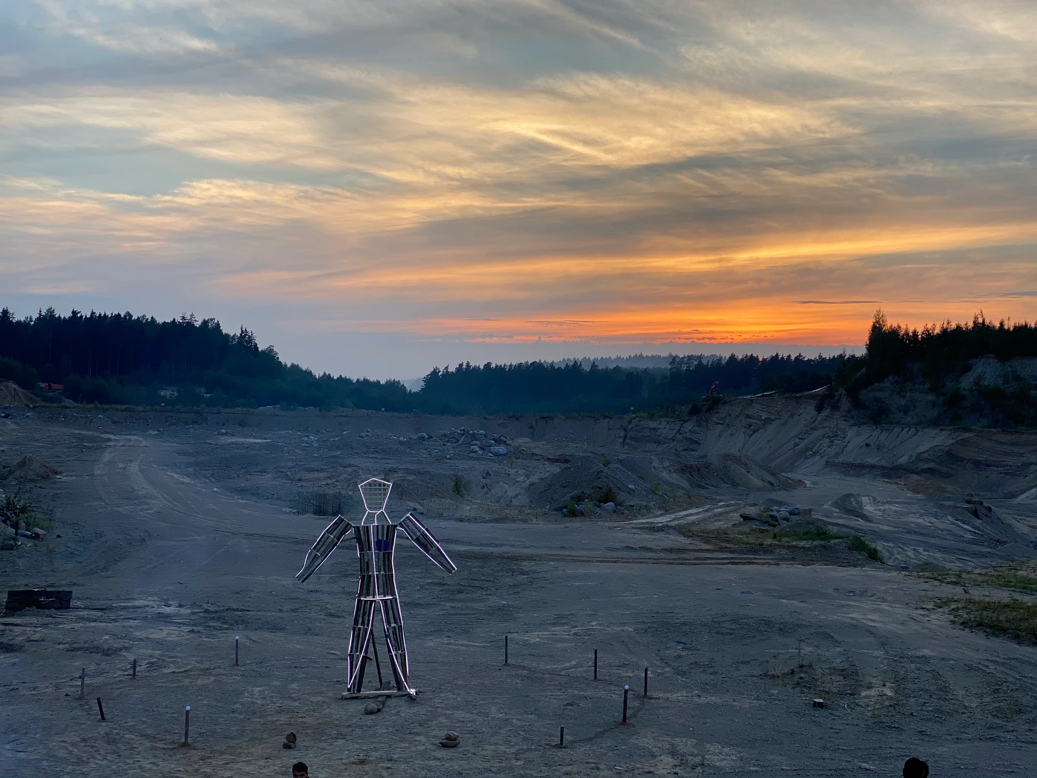 A wooden statue of a humanoid figure adorned with LED lights stands against a barren background with a beautiful orange sunset
