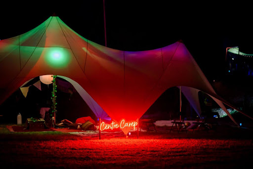 A large white marquee sits glowing in the dark night with multi coloured lights and a light up sign saying Centre Camp in front of it