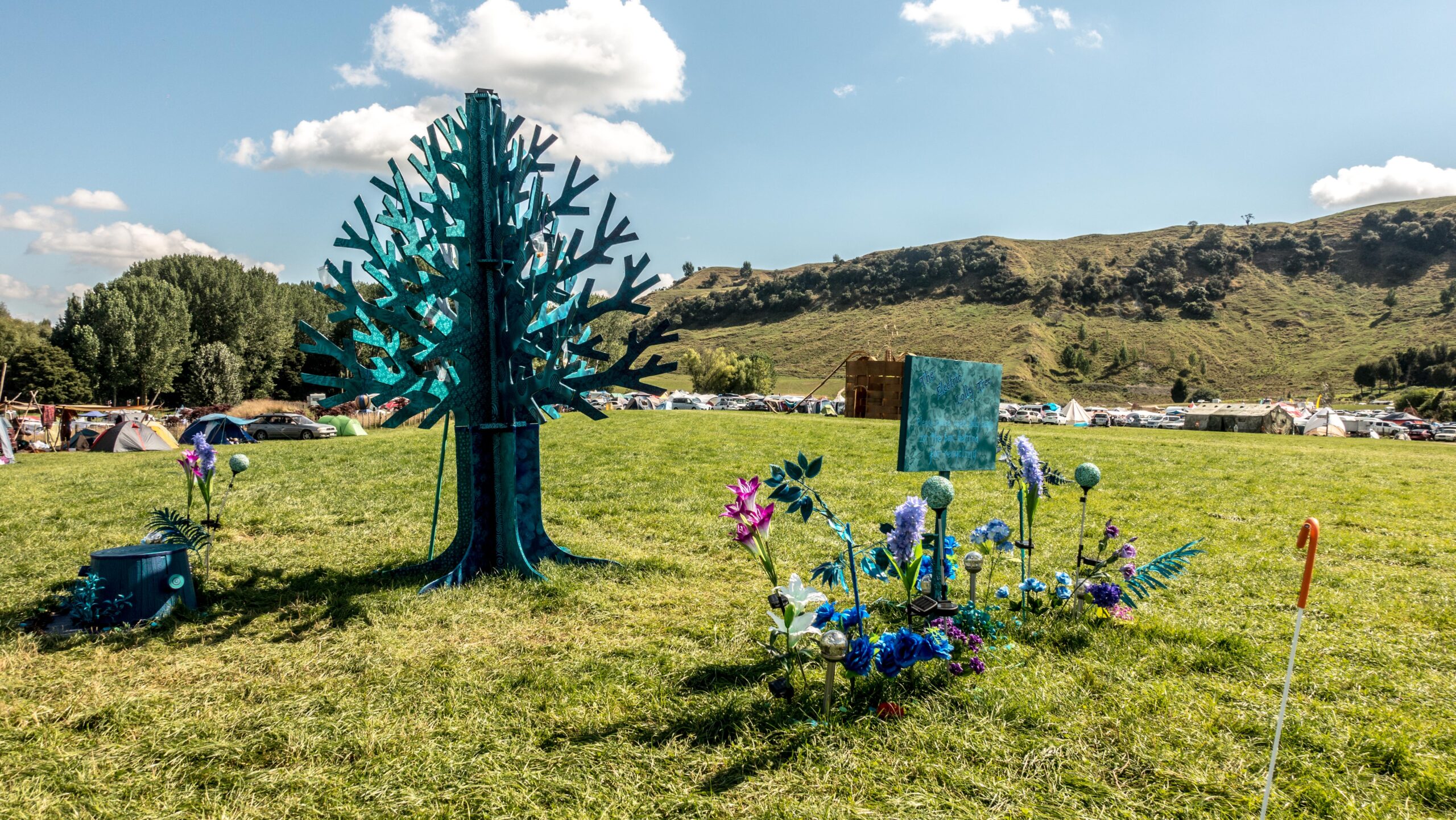 A bluish green wooden tree stands tall in front of colourful wooden flowers with tents and a lush green paddock in the background