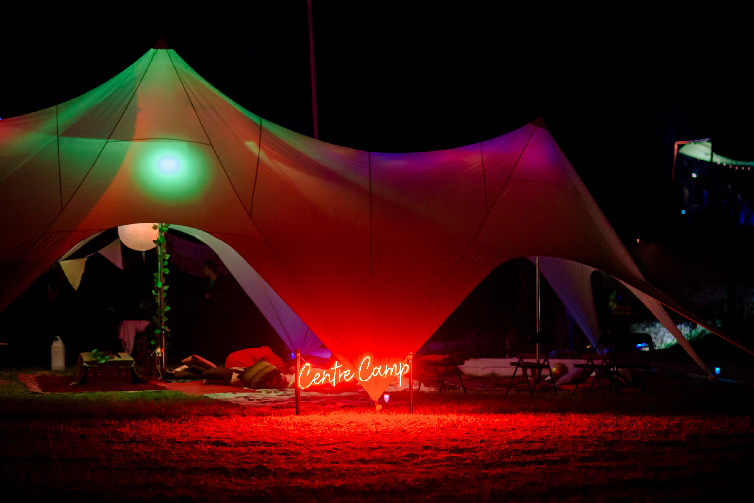 A white marquee lit by bright multi-coloured lights sits against the night, with a red LED sign saying Centre Camp in front of it