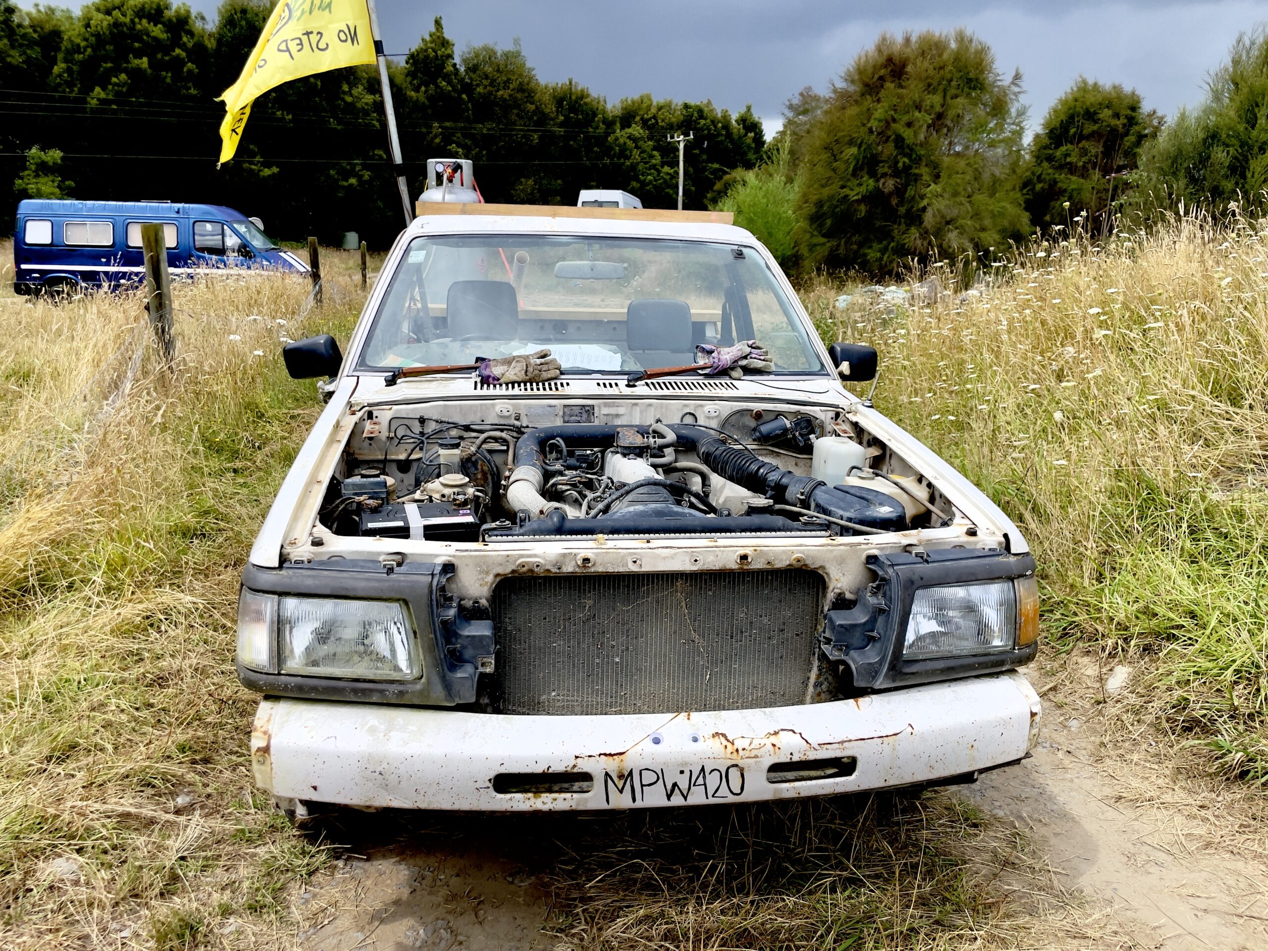 A dusty, rusty white car with no bonnet sits on a dirt road in a paddock