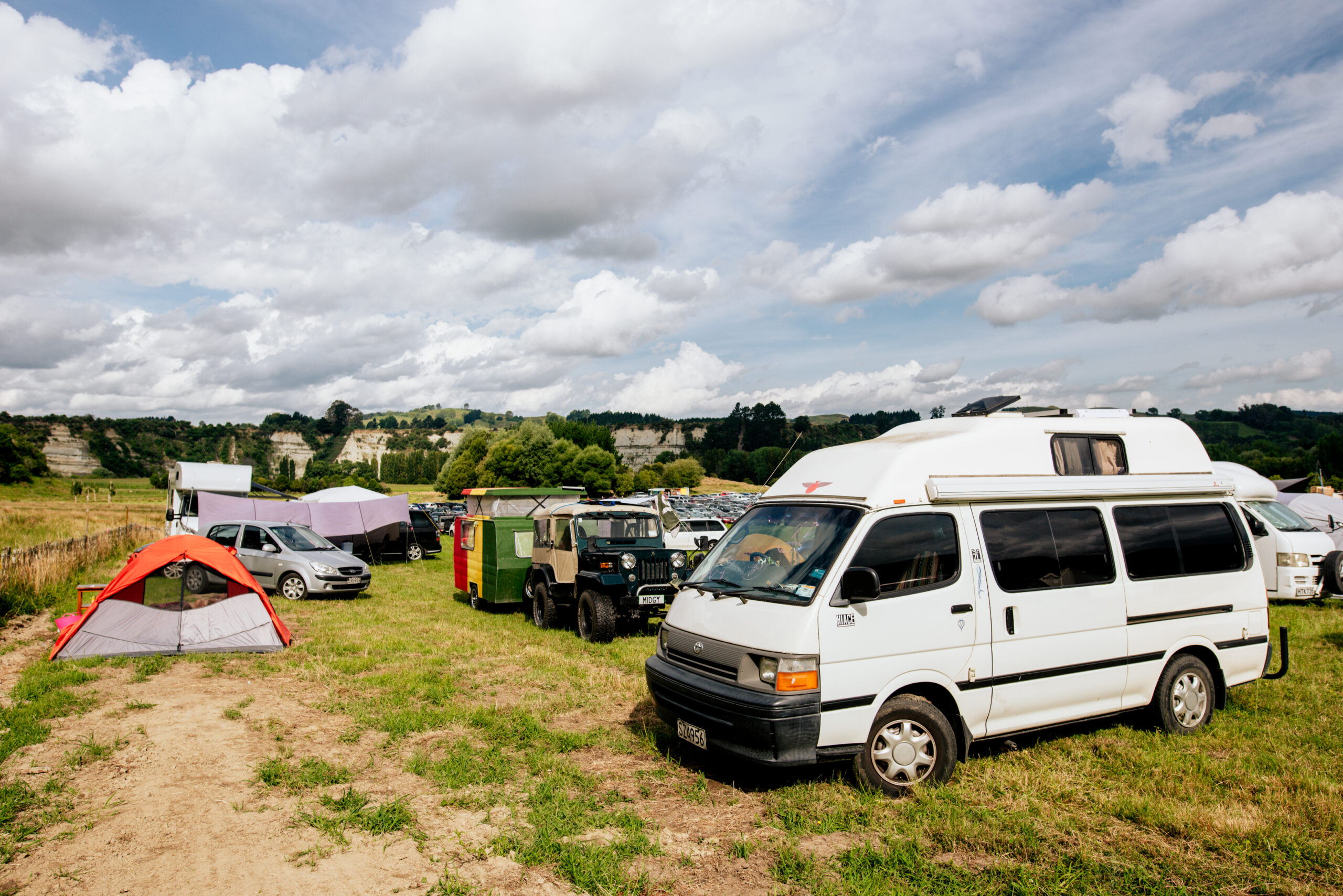 Camper vans and tents sit on a green paddock, a light cloud sky in the background