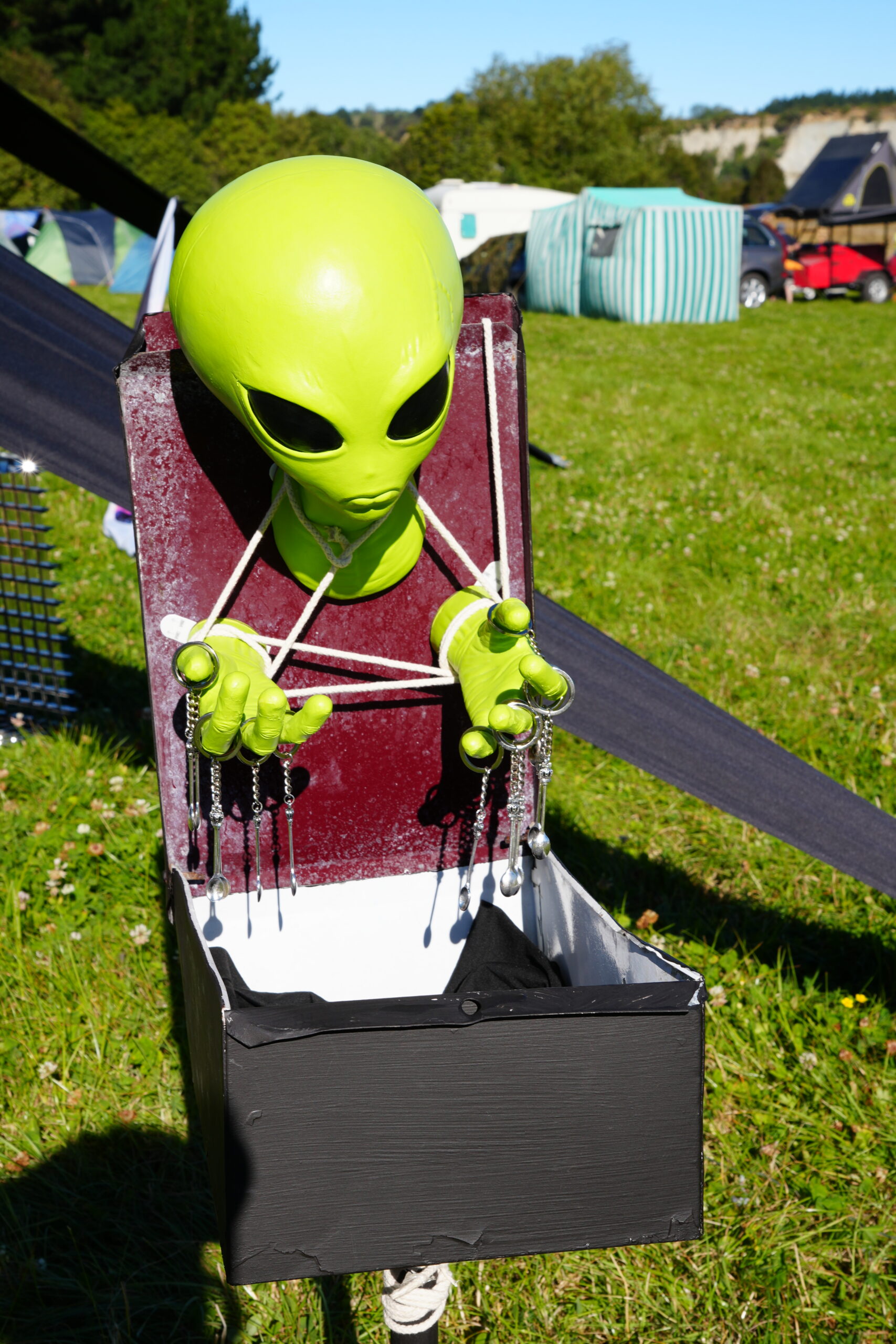 Artwork.maxvolk.2025.cosmicrejex5 A green alien statue art work with a box hanging around their neck