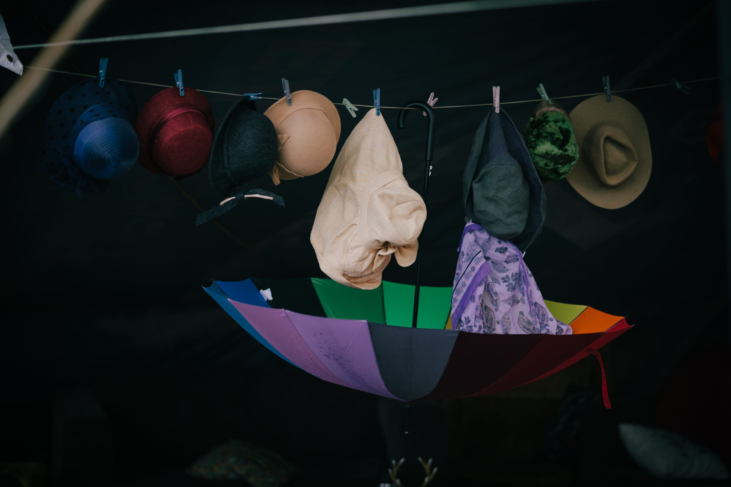 An array of hats hangs from a clothes line, with an upside down rainbow umbrella in the centre