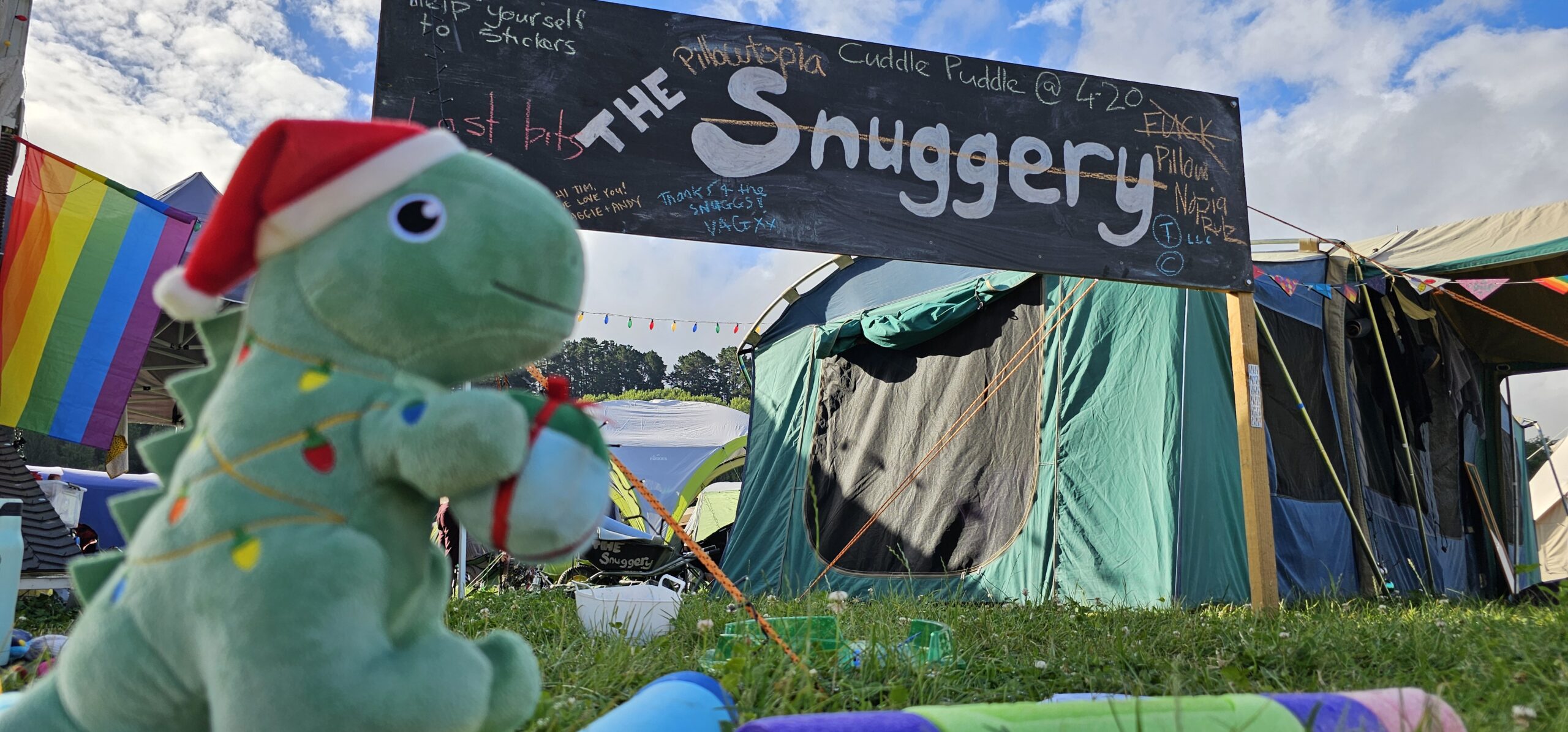 20240127_182958-snuggery-Keegan Durant A green stuffed toy t-rex holds a small gift, standing in front of a tent and a sign that reads 'the snuggery'