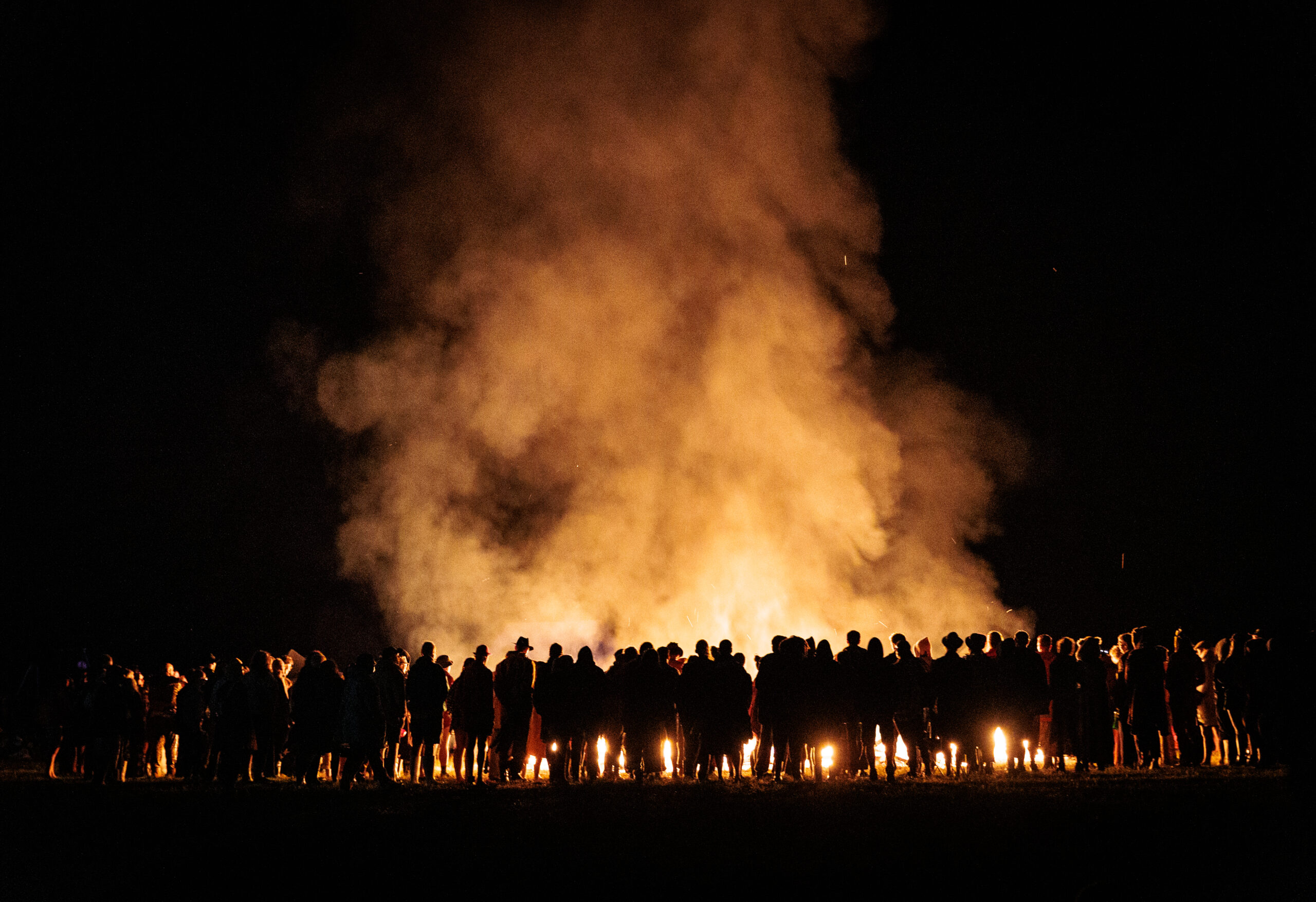 A group of people stands silhouetted in front of an enormous fire