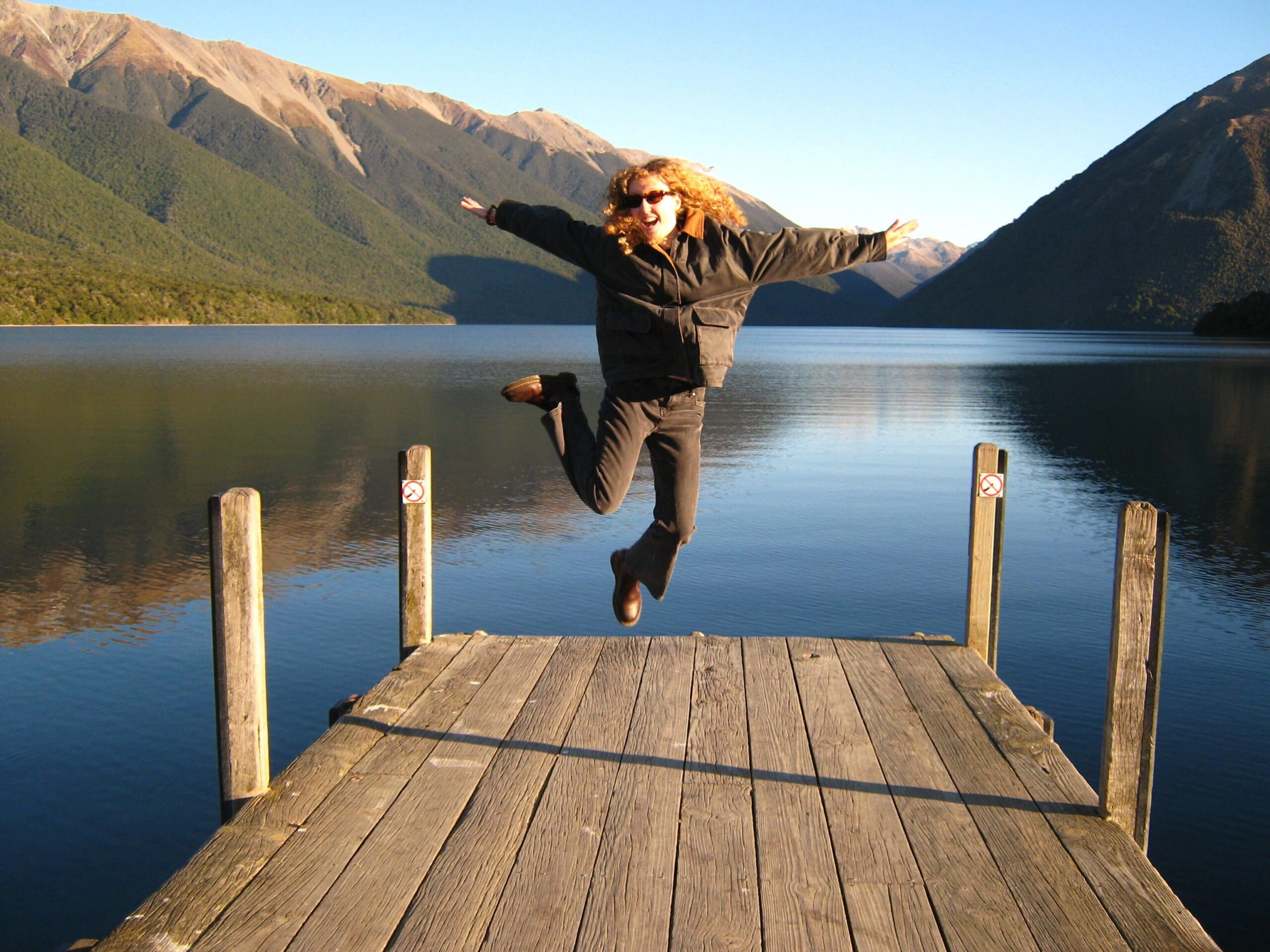 Image from iOS New blog writer Lilly is jumping at the end of a wharf that juts out into a beautiful lake with mountains reflected on its surface