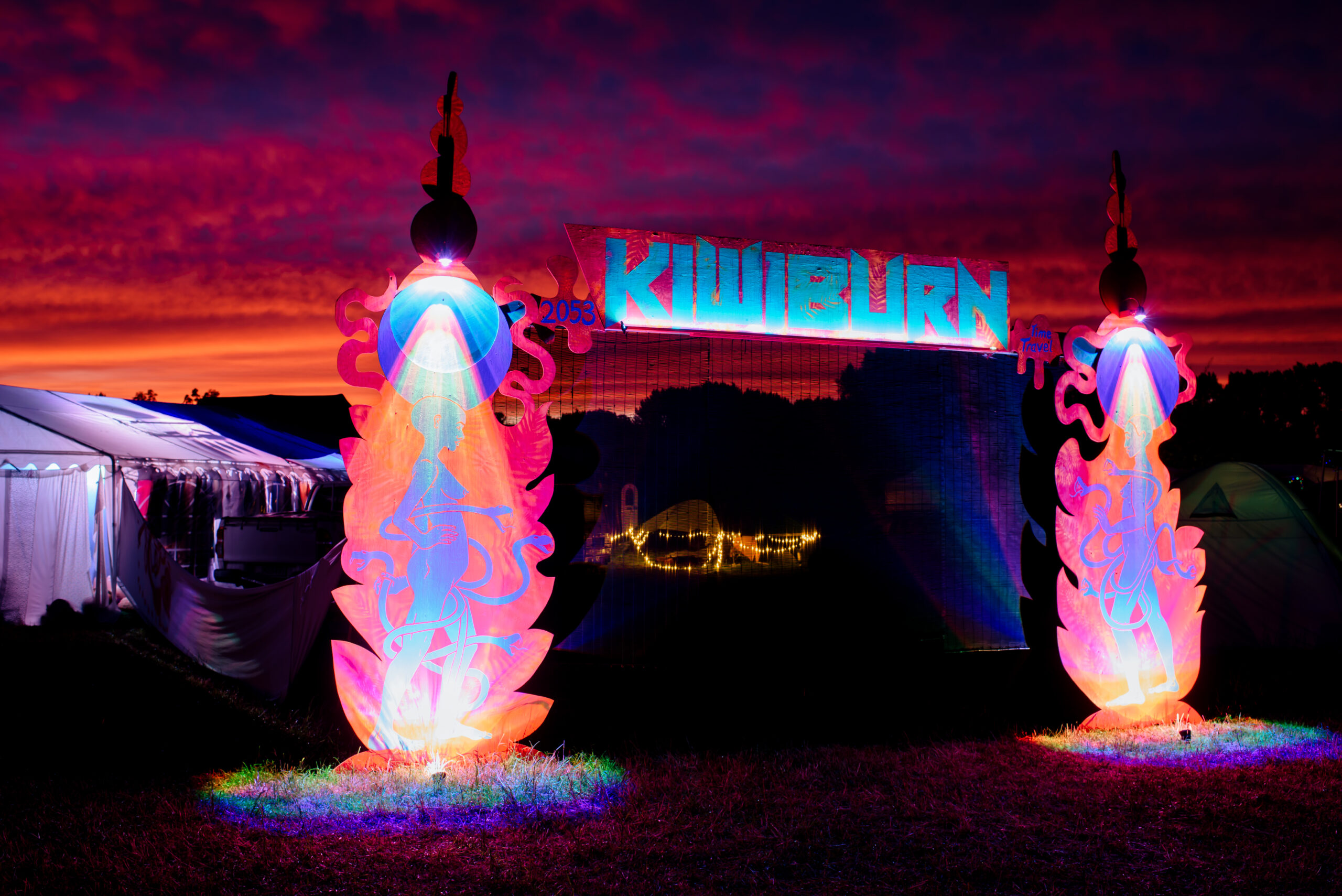 Nabulen_Kiwiburn23-67 a glowing sign saying kiwiburn sits above two figures with the sun setting in the background