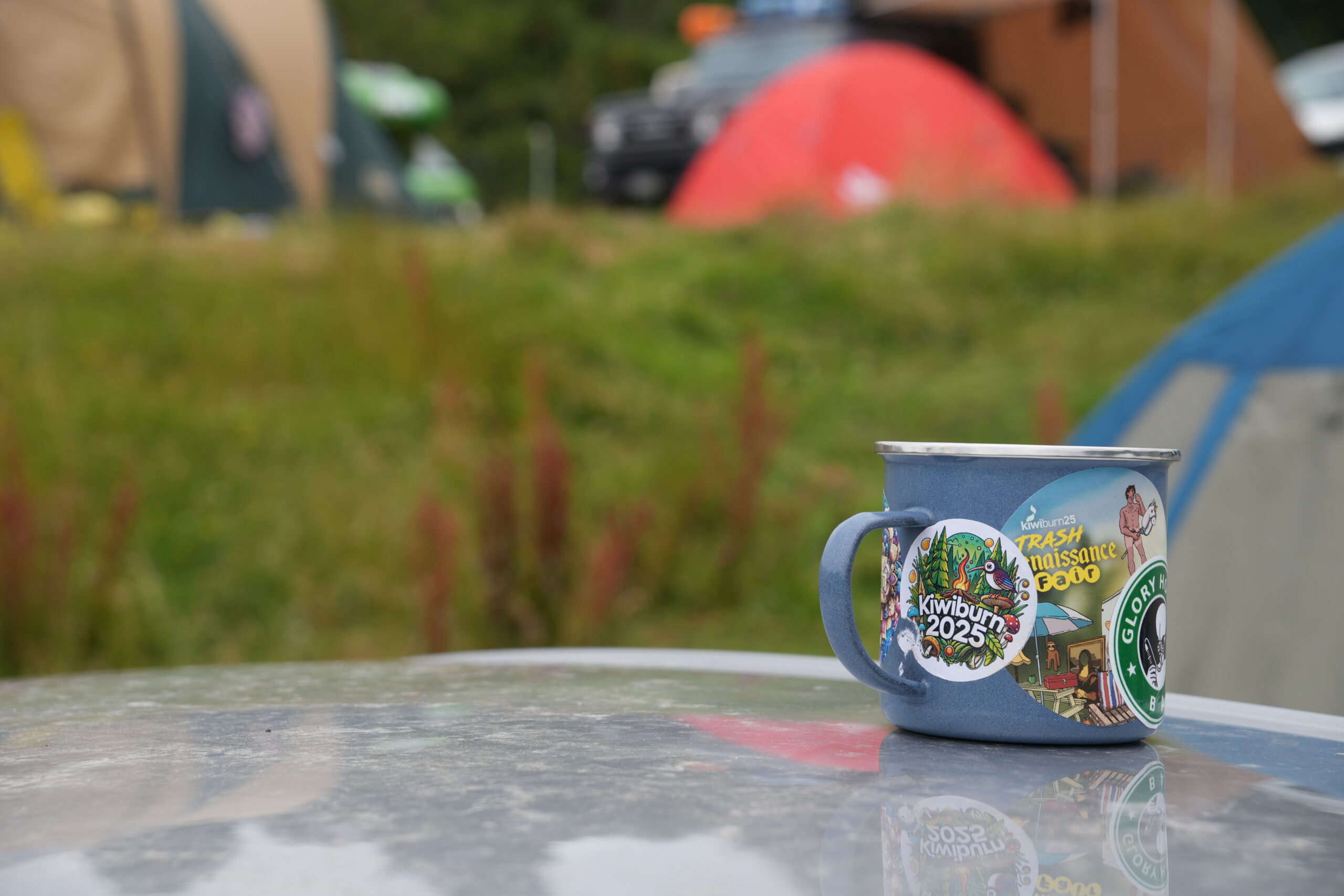 vessel-mug.maxvolk.2025.two A camp mug covered in stickers sits on a table with blurry tents in the background