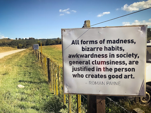A sign on a green field which says "All forms of madness, bizarre habits, awkwardness in society, general clumsiness, are justified in the the person who creates good art. - Roman Payne"